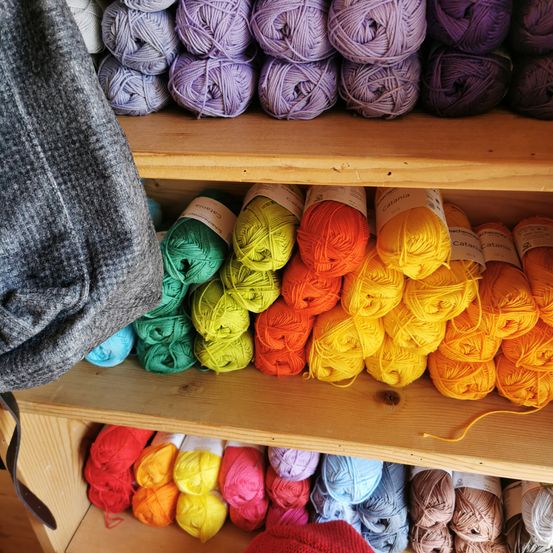 A wooden shelf displays a variety of yarns in different colors, with some shades of purple on top and a mix of vibrant colors below.