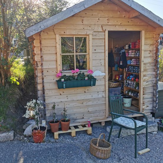 A small wooden cabin with a window and flowers in a planter box. Inside, there are shelves with yarns, and a chair with a cushion. Outside, there are potted plants and a basket.