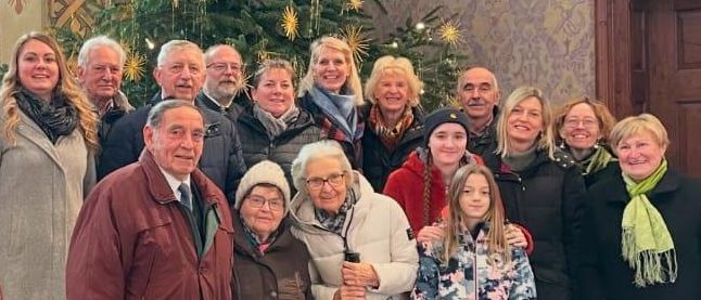Eine Gruppe von Menschen, wahrscheinlich Familie, versammelt sich vor einem geschmückten Weihnachtsbaum. Sie stehen auf einem gefliesten Boden mit einer Holztür hinter ihnen.