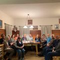 Eine Gruppe von Erwachsenen, meist ältere Menschen, sitzen in einem Esszimmer mit Tisch, Gläsern und Tellern. Der Raum hat Holzwände und Vorhänge.