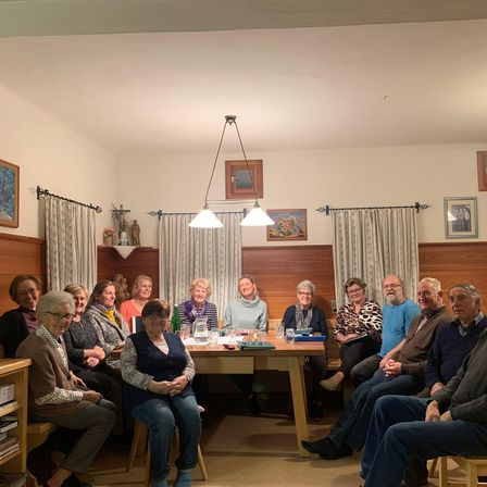 Eine Gruppe von Erwachsenen, meist ältere Menschen, sitzen in einem Esszimmer mit Tisch, Gläsern und Tellern. Der Raum hat Holzwände und Vorhänge.