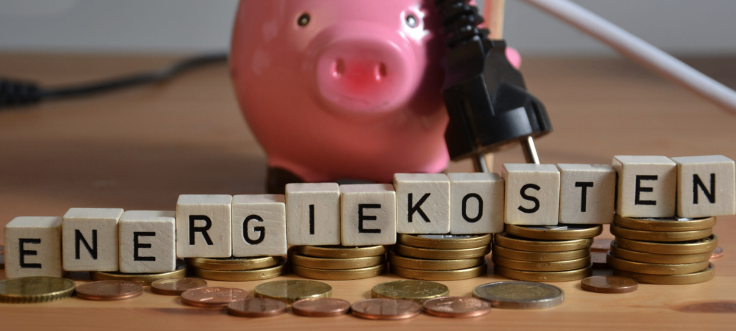 Eine Sparschwein mit Stecker und Stapeln von Münzen und Würfeln, die Energiekosten spellen.