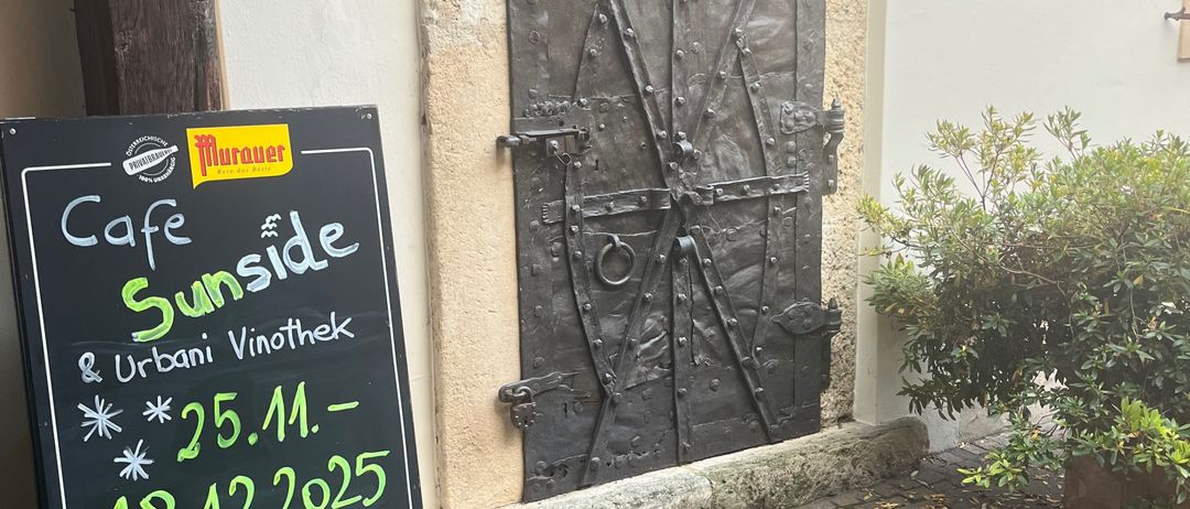 Ein Schwarzes Brett für Cafe Sunside und Urbani Vinothek steht neben einer Metalltür mit Griff und einer Pflanze auf dem Boden. Das Datum ist auf dem Schild geschrieben.
