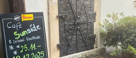 Ein Schwarzes Brett für Cafe Sunside und Urbani Vinothek steht neben einer Metalltür mit Griff und einer Pflanze auf dem Boden. Das Datum ist auf dem Schild geschrieben.