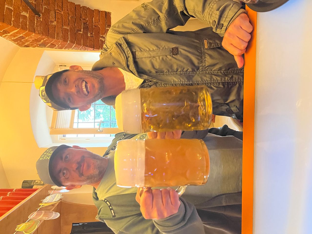 Zwei Männer in grünen Uniformen und Mützen lächeln und halten große Gläser Bier hoch. Der Mann auf der linken Seite trägt eine Mütze mit einem Logo. Im Hintergrund ist eine Ziegelmauer und ein Fenster zu sehen.