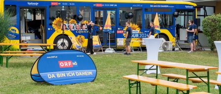 Ein blau-gelber Bus mit ORF-Logo steht draußen. Eine Person steht in der Nähe, mit zwei weiteren bei einem Tisch und Bänken.