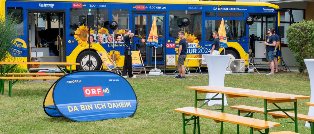 Ein blau-gelber Bus mit ORF-Logo steht draußen. Eine Person steht in der Nähe, mit zwei weiteren bei einem Tisch und Bänken.