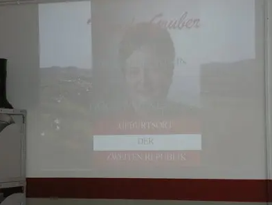 Eine Beamerleinwand zeigt eine Folie mit einem Foto einer Frau und den Text 'DEUBURTSORT' und 'DEK' darunter. Darunter steht der Text 'ZWEITEN REPUBLIK'.