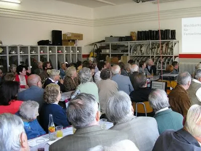 Eine Gruppe älterer Erwachsener, einige mit Hüten, sitzen an Tischen in einem Raum. Einige von ihnen betrachten eine Tafel mit Text.