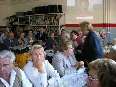 Eine Gruppe älterer Menschen sitzt in einem Raum an einem Tisch, einige essen und trinken, andere hören einer Präsentation auf einer Projektionsleinwand zu.