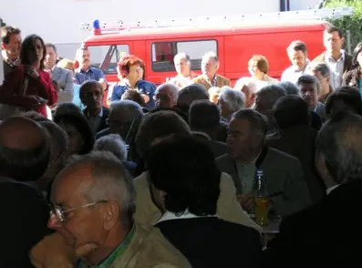 Eine große Menschenmenge versammelt sich draußen, einige sitzen an Tischen, andere stehen. Ein roter Feuerwehrwagen ist hinter ihnen geparkt.