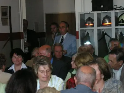 Eine Gruppe von Erwachsenen, sowohl Männer als auch Frauen, sind in einem Raum versammelt. Einige tragen Brillen, Krawatten und formelle Kleidung. Ein Mann in der Mitte hält einen Becher. Dahinter befindet sich ein Regal mit Helmen, einer Lampe und einem Lautsprecher.