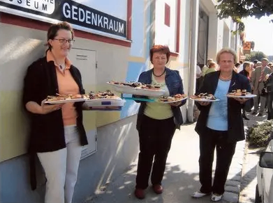 Drei Frauen stehen nebeneinander und halten Tabletts mit Essen vor einem Gebäude mit dem Schild Gedenkraum.
