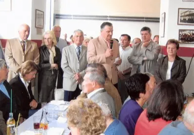 Eine Gruppe formell gekleideter Männer und Frauen steht und sitzt in einem Raum. Ein Mann steht vor einem Mikrofon und scheint zu sprechen.