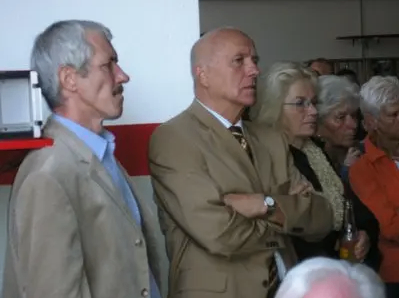 Zwei Männer und eine Gruppe von Menschen stehen in einem Raum zusammen, wobei der Mann auf der linken Seite den Mann in der Mitte ansieht.