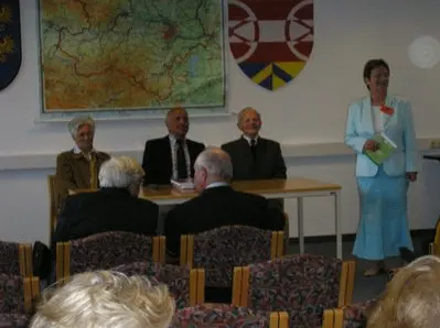 Vier Männer und eine Frau in formeller Kleidung sitzen an einem Tisch in einem Konferenzraum, mit einer Frau in einem blauen Kleid, die steht und ein Buch hält. Hinter ihnen befinden sich mehrere Stühle, und eine Karte und ein Schild hängen an der Wand hinter dem Tisch.