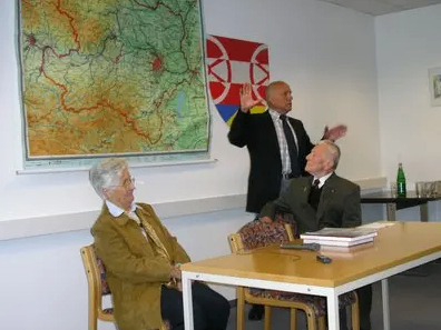 Drei Männer in Anzügen in einem Besprechungsraum, einer steht und gestikuliert, während die anderen beiden in Stühlen sitzen. Eine Karte und eine Flagge sind an der Wand.