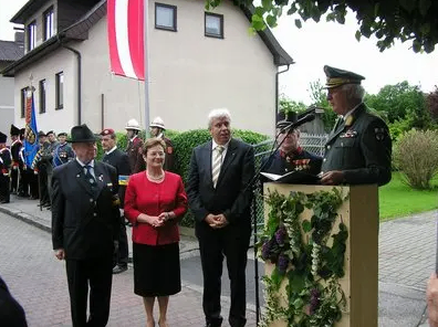 Ein Mann in Uniform hält eine Rede an einem Podium, wobei eine Frau in einem roten Blazer und ein Mann in einem schwarzen Anzug neben ihm stehen. Dahinter stehen eine Flagge und mehrere Menschen in Uniform.