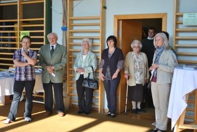 Eine Gruppe von Menschen, einschließlich älterer Erwachsener, steht in einem Raum mit Tischen und Stühlen, wahrscheinlich bei einer Veranstaltung.