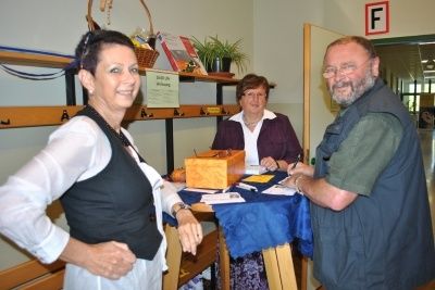 Drei Personen stehen lächelnd neben einem Tisch mit blauer Tischdecke. Ein Holzkasten und Papiere liegen auf dem Tisch. Ein Regal mit Büchern und einem Korb steht hinter ihnen.
