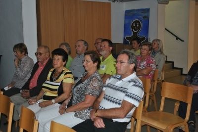 Eine Gruppe von Erwachsenen, darunter Männer und Frauen, sitzen in Stühlen in einem Raum, wahrscheinlich an einer Besprechung teilnehmend.