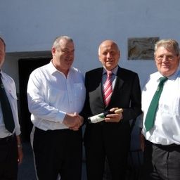 Vier Männer in formeller Kleidung stehen vor einem Gebäude. Der Mann in der Mitte hält eine Flasche und scheint sie dem Mann rechts von ihm zu präsentieren.