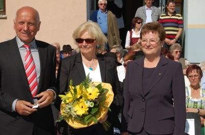 Drei Menschen stehen eng beieinander, zwei Frauen halten einen Strauß gelber Blumen und ein Mann hält ein Stück Papier. Dahinter steht eine Menschenmenge, einige tragen Brillen und Hüte.