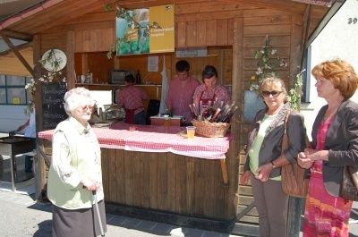 Eine Frau mit einem Gehstock steht hinter einem Tresen mit einer rot-weiß karierten Tischdecke. Ein Korb mit Gegenständen steht auf dem Tresen. Drei Frauen stehen und unterhalten sich vor dem Tresen.