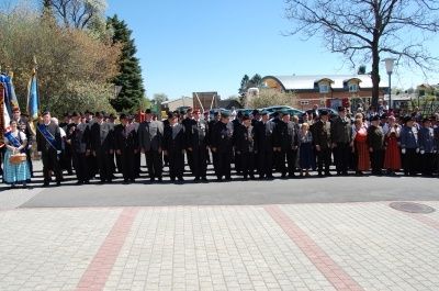 Eine große Gruppe von Menschen in formeller Kleidung steht in einer Reihe draußen, einige halten Fahnen, mit Bäumen und Gebäuden im Hintergrund.