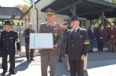 Ein Militäroffizier überreicht einem anderen Offizier eine Auszeichnung im Freien. Hinter ihnen steht eine Gruppe von Menschen und beobachtet.