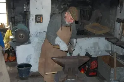 Ein älterer Mann in einer Werkstatt, der eine Mütze und eine Schürze trägt, hält einen Hammer und arbeitet an einem Amboss. Ein Eimer und ein Rad sind in der Nähe.