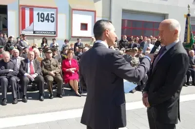 Zwei Männer in Anzügen und Krawatten sprechen in Mikrofone vor einer Menschenmenge. Ein Mann hält ein Mikrofon in der rechten Hand. Dahinter steht ein großes Schild mit dem Jahr 1945.