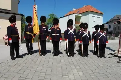 Eine Gruppe von Soldaten in Uniform steht in einer Reihe auf einem Ziegelpflaster. Sie halten Flaggen und Gewehre. Hinter ihnen stehen Gebäude, von denen eines ein rotes Dach hat.