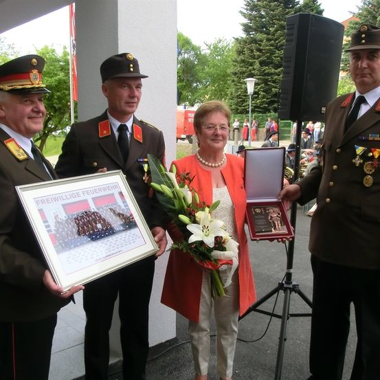 Eine Frau erhält eine Auszeichnung, während sie zwischen zwei Männern steht. Die Frau trägt eine Brille und hält einen Blumenstrauß. Die Männer sind in Militäruniformen gekleidet.