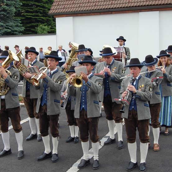Eine Gruppe von Männern in traditioneller bayerischer Kleidung spielt Blech- und Holzblasinstrumente. Sie stehen vor einem weißen Gebäude, mit Bäumen im Hintergrund.