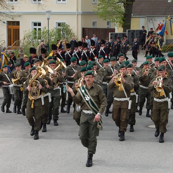 Eine Marschkapelle in grünen Uniformen und schwarzen Stiefeln, die Blechblasinstrumente spielt, zieht durch einen Stadtplatz.