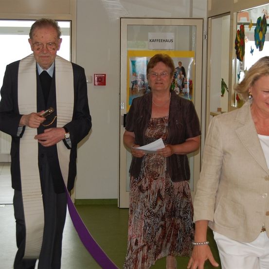 Drei Personen stehen in einem Raum, und eine Frau hält ein Papier in der Hand. Ein Mann trägt einen schwarzen Mantel und eine weiße Stola, und eine Frau trägt ein braunes Kleid. Hinter ihnen befindet sich eine Glastür mit der Aufschrift 'KAFFEEHAUS'.