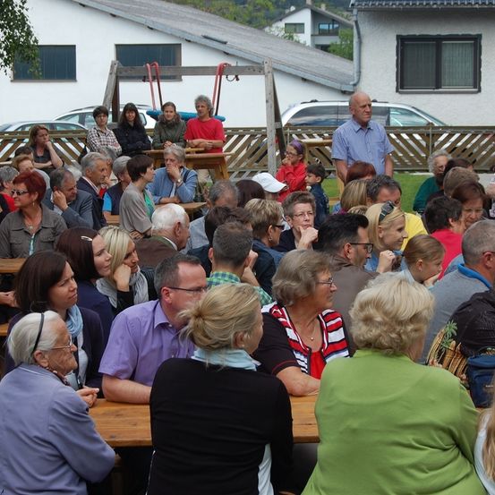 Eine Menschenmenge, hauptsächlich Frauen, sitzt an Tischen im Freien. Ein Mann steht auf dem Rasen und redet. Ein paar Kinder sind in der Nähe. Im Hintergrund befinden sich Häuser, ein Zaun und geparkte Autos.