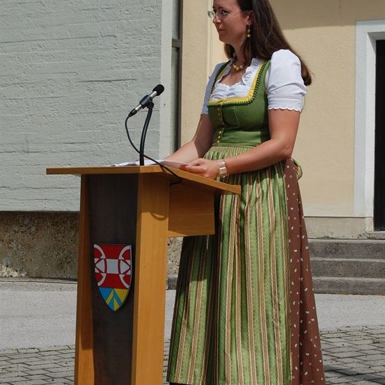 Eine Frau in traditioneller Kleidung steht vor einem Podest mit einem Mikrofon vor einem Gebäude.