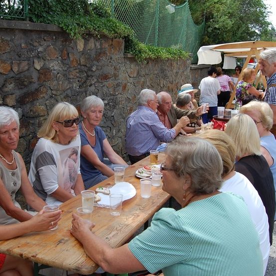 Eine Gruppe älterer Menschen sitzt um einen Holztisch herum, wahrscheinlich beim Picknick. Auf dem Tisch stehen Teller, Gläser und Essen. Hinter ihnen steht eine Steinmauer mit Pflanzen, und ein Mensch steht vor ihnen.