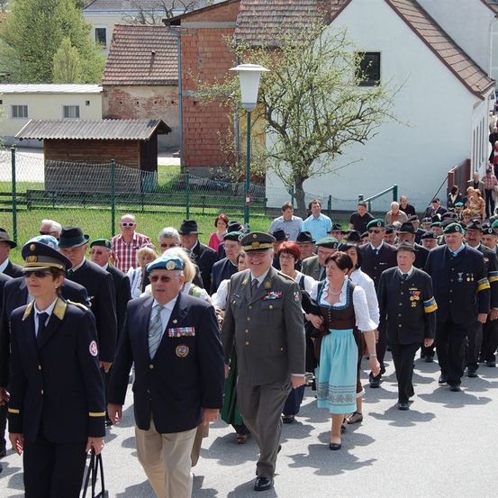 Eine Gruppe von Menschen in Uniformen und Hüten, die auf einer Straße mit Gebäuden und einem Zaun im Hintergrund gehen.