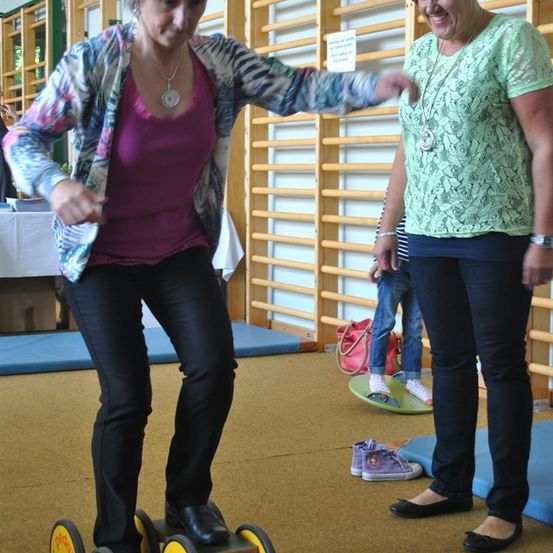 Zwei Frauen sind in einem Raum mit Holzregalen im Hintergrund. Eine von ihnen balanciert auf einem Gerät mit Rädern.