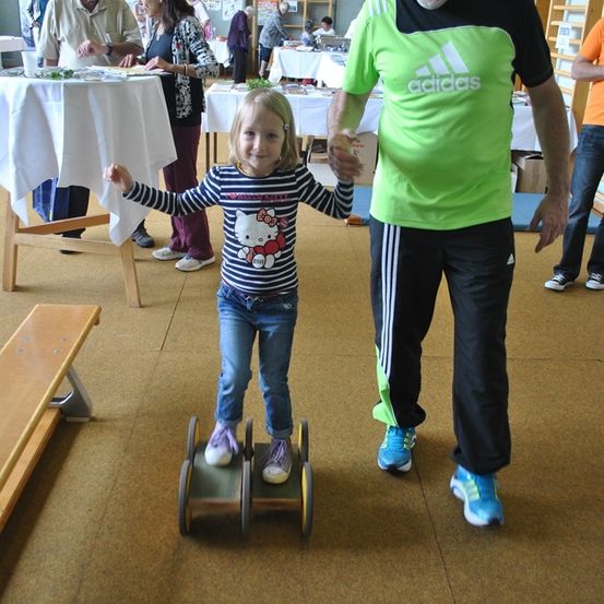 Ein Mädchen läuft auf einem kleinen Fahrzeug und hält die Hände eines älteren Mannes. Dahinter stehen Leute in der Nähe von Tischen.