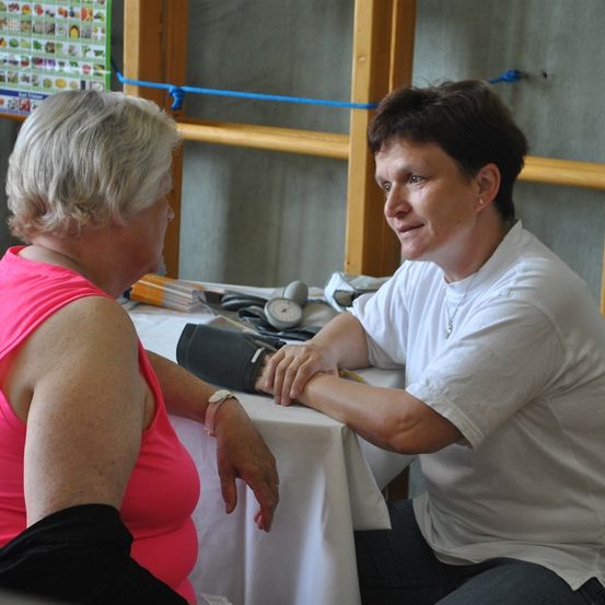 Eine Frau in einem rosa Oberteil unterhält sich mit einer Frau in einem weißen Oberteil. Die Frau in Weiß hält ein Blutdruckmessgerät und lächelt.