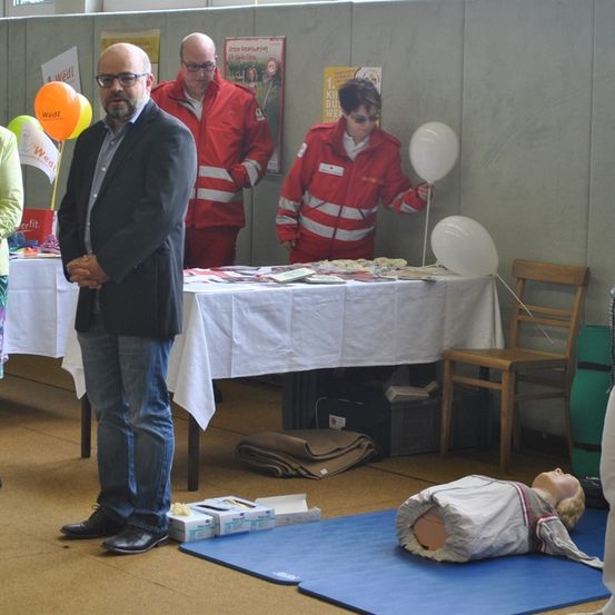 Ein Mann steht neben einem Tisch mit Materialien und einer Frau, die einen Ballon hält. Eine andere Frau steht hinter dem Tisch mit einer Puppe, die auf dem Boden liegt.