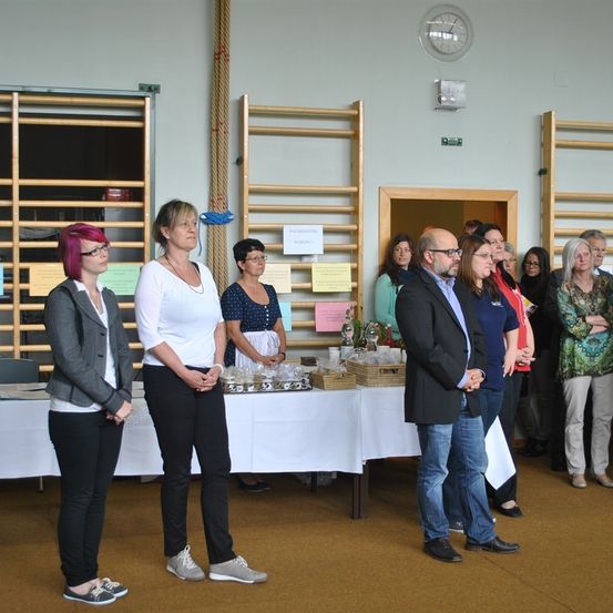 Eine Gruppe von Menschen, darunter Frauen in formeller Kleidung, steht in einem Raum mit einem Tisch vor ihnen. Ein Mann steht neben ihnen und eine Frau steht hinter ihnen. Eine Frau mit rosa Haaren steht vor dem Tisch, mit einer Uhr an der Wand darüber.