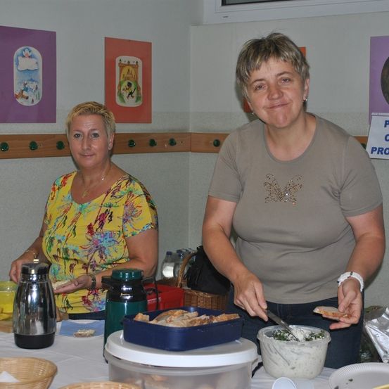 Zwei Frauen stehen lächelnd hinter einem Tisch. Eine von ihnen hält ein Messer und Brot, während die andere ein Stück Brot in der Hand hat. Sie befinden sich in einem Raum mit Gemälden an der Wand.