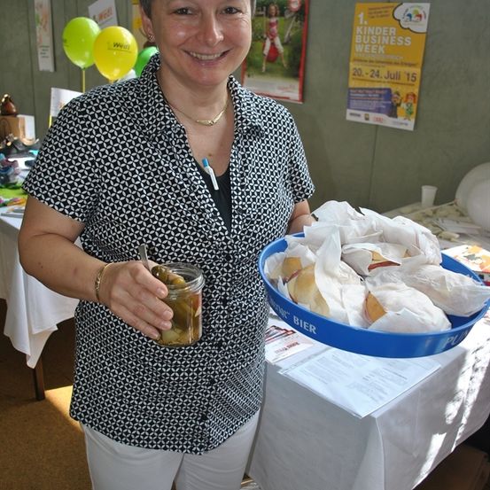 Eine lächelnde Frau mit einer Halskette steht vor einem Tisch und hält ein Glas mit Deckel und Löffel. Dahinter befindet sich ein Tisch mit weißer Tischdecke und eine blaue Schüssel mit Essen. An der Wand hängen auch Luftballons, ein Plakat und ein Schild.