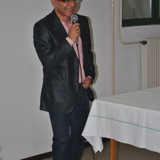 Ein Mann in einem schwarzen Anzug, der ein Mikrofon hält, spricht zu einer Menschenmenge in einem Raum mit einer weißen Wand und einem Tisch mit einer weißen Tischdecke.