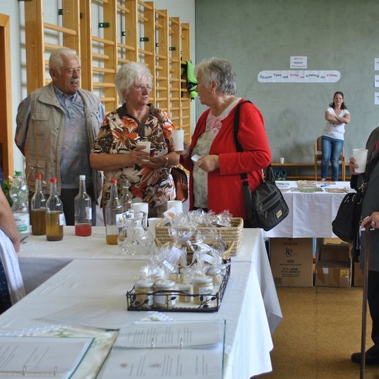 In einem Raum mit Holzregalen stehen vier ältere Menschen um einen Tisch mit Flaschen und Körben. Sie tragen Freizeitkleidung und scheinen miteinander zu sprechen.